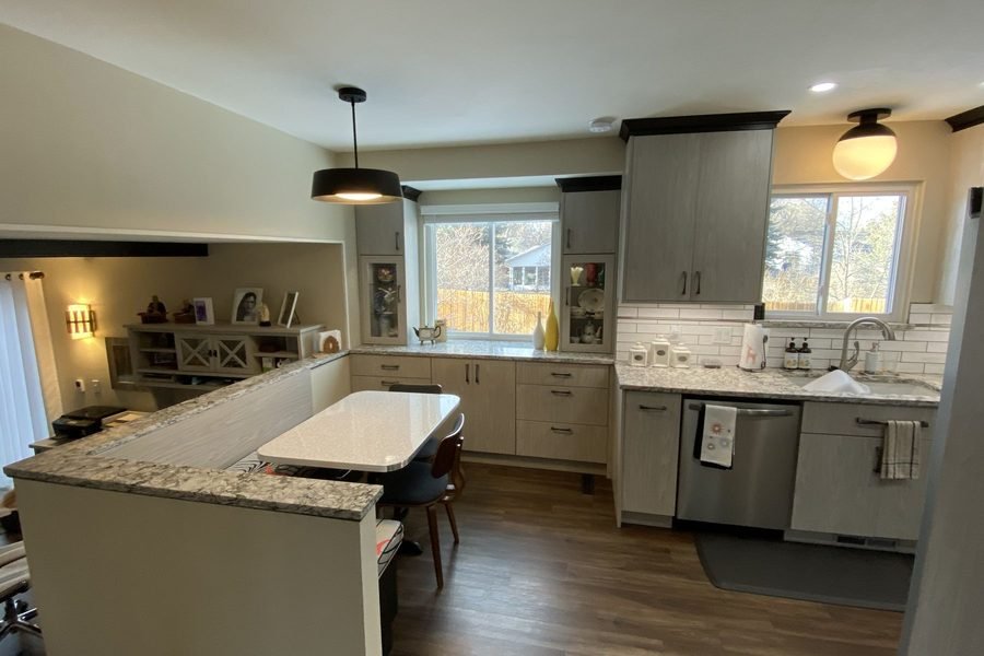 A finished kitchen with light greige cabinets, granite counters, a raised bar peninsula with seating, a white subway tile backsplash, a stainless dishwasher and a black drum pendant light