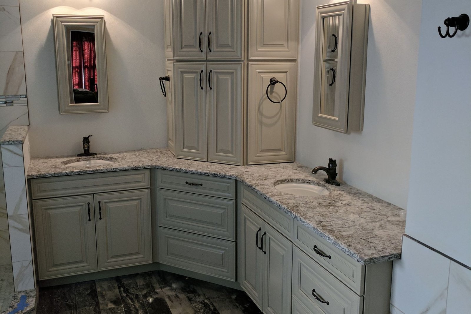 A finished bathroom with cream glazed cabinets set into a corner, a tall storage tower between two sinks, granite counters, oil rubbed bronze faucets and sconces, and a dark wood-look plank floor