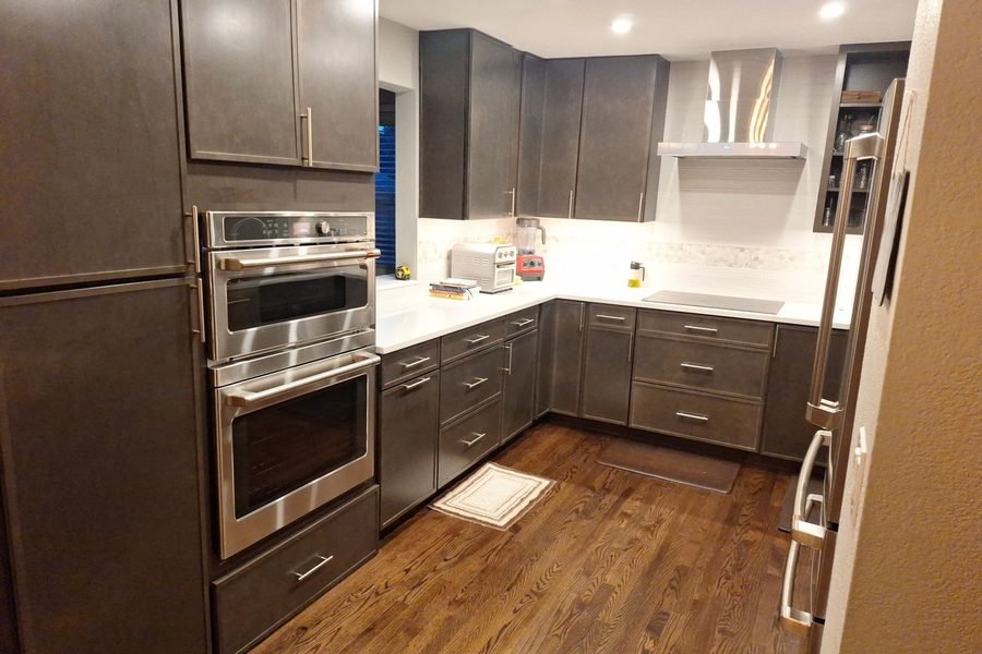 A finished kitchen with grey brown flat panel slab cabinets, white counters, stainless double wall ovens built into a tall cabinet run, a stainless chimney hood and a hardwood floor