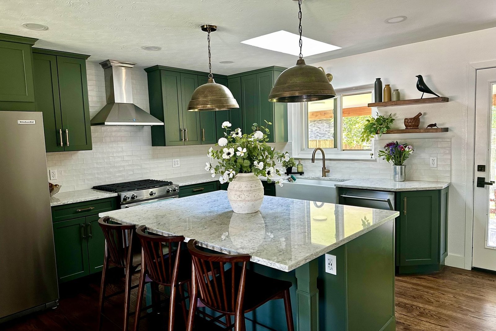 A finished kitchen with deep green shaker cabinets, a white subway tile backsplash, a stainless chimney hood over the range, two brass dome pendant lights and a granite island with three wooden stools