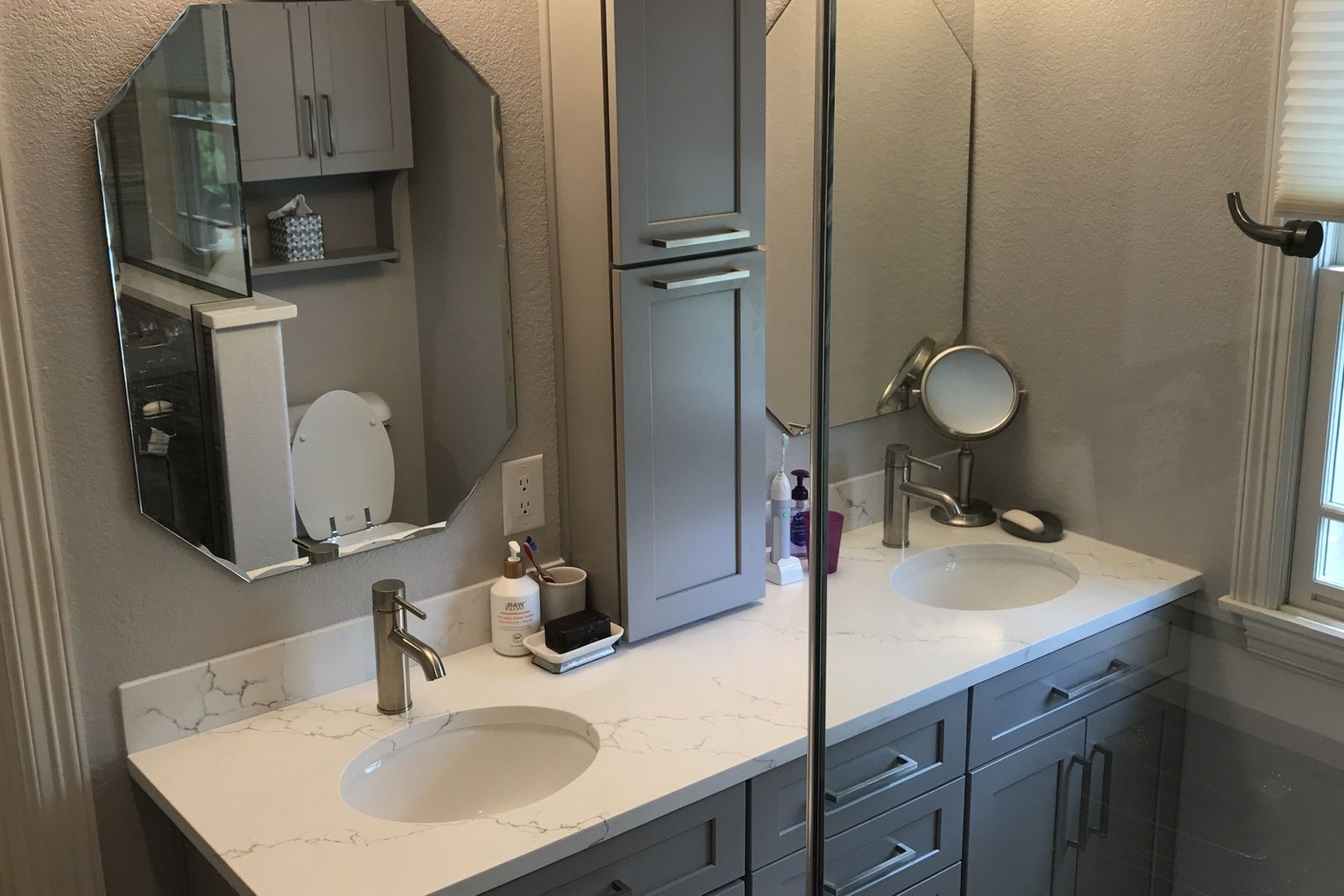 A finished bathroom vanity in grey cabinetry with two oval undermount sinks, a white quartz counter with grey veining, a tall storage tower between the sinks and an octagonal mirror