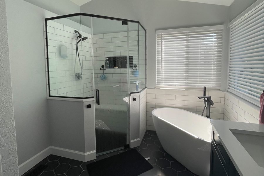 A finished bathroom under a sloped ceiling, with a glass neo angle shower in white subway tile, a freestanding tub in the window corner, large black hexagon floor tile and matte black fittings