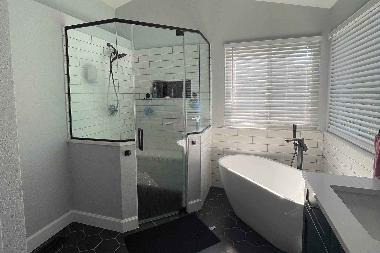 A finished bathroom under a sloped ceiling, with a glass neo angle shower in white subway tile, a freestanding tub in the window corner, large black hexagon floor tile and matte black fittings
