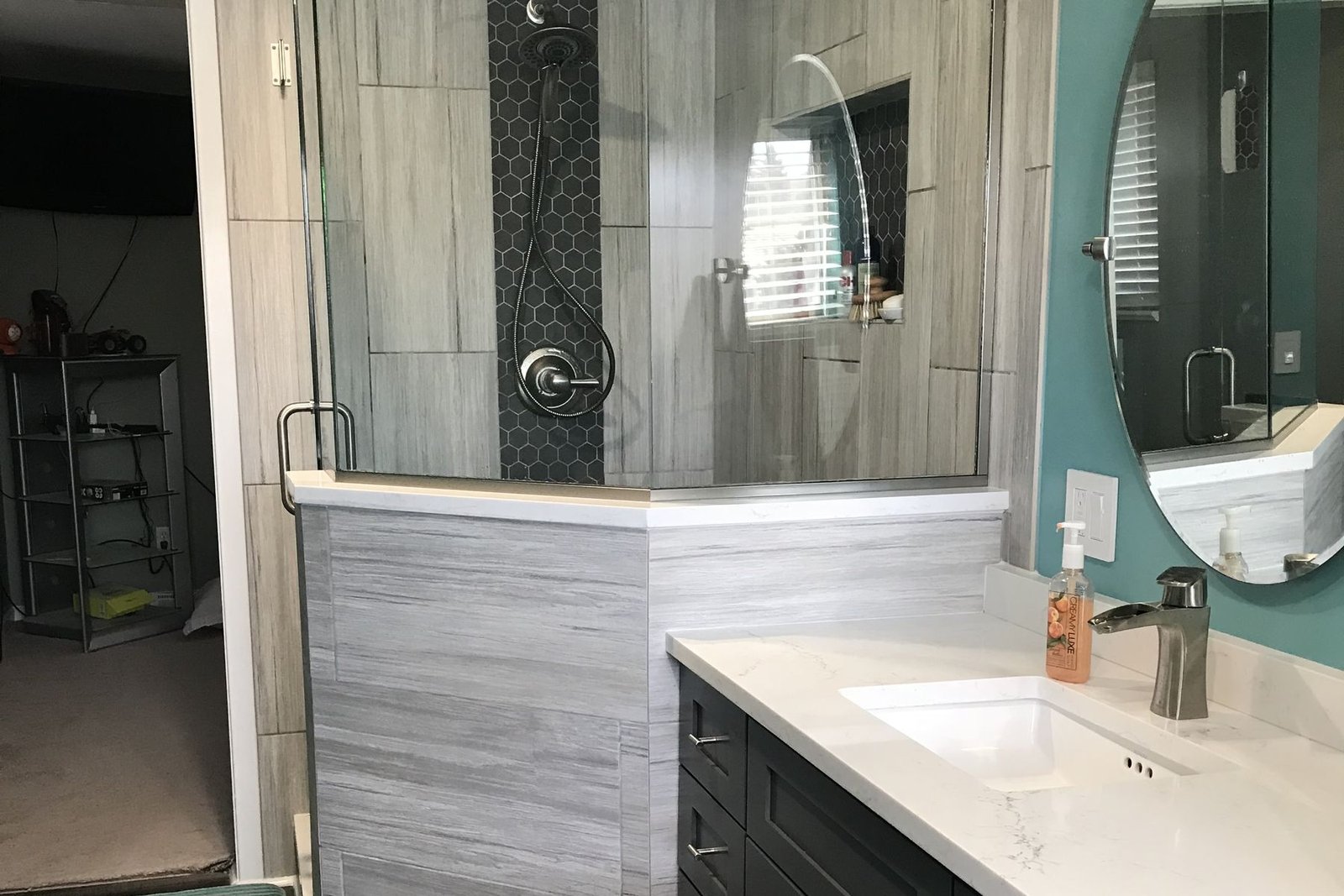 A finished bathroom with a corner shower behind a glass panel and a low tiled half wall, a vertical black hexagon mosaic stripe running floor to ceiling behind the shower fixtures, grey linear-grain wall tile, and a dark shaker vanity with a white stone top and a brushed steel faucet