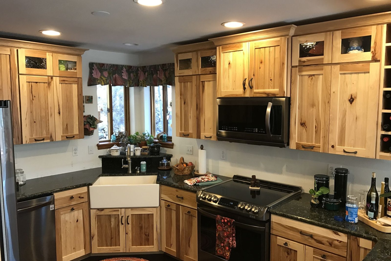 A finished kitchen with knotty hickory cabinets, dark granite counters, a white apron front farmhouse sink under a run of windows, a black range and glass fronted upper cabinets