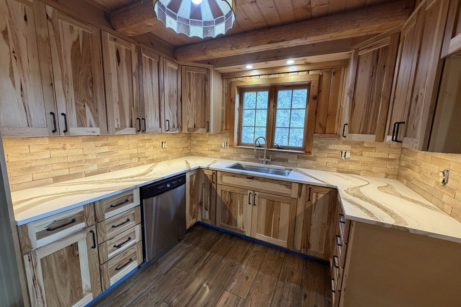 A finished U-shaped kitchen under log beams and a stained-glass pendant: hickory shaker cabinets with black pulls, white counters with gold and brown veining, a stainless sink under the window, a stainless dishwasher and a brick-style tile backsplash