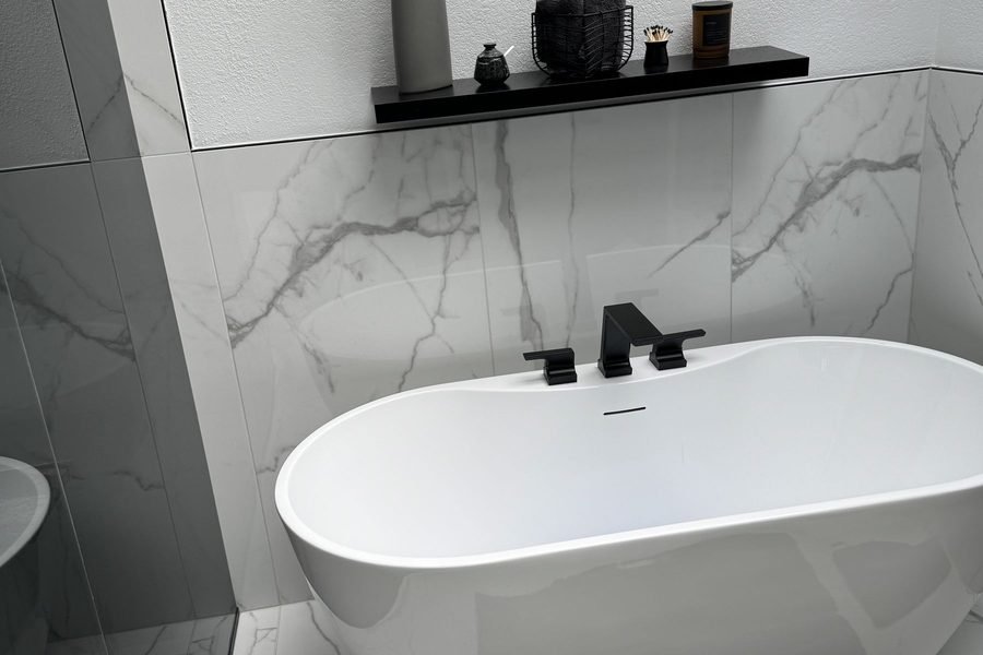 A finished bathroom in large format white marble look porcelain on the walls and floor, with a freestanding oval tub, a matte black tub filler and a black floating shelf