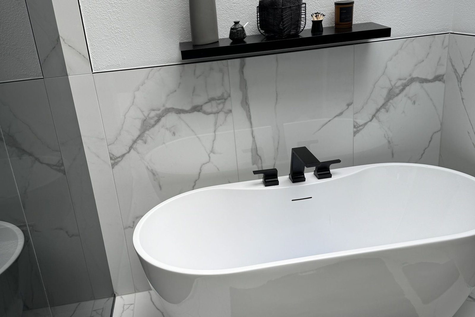 A finished bathroom in large format white marble look porcelain on the walls and floor, with a freestanding oval tub, a matte black tub filler and a black floating shelf