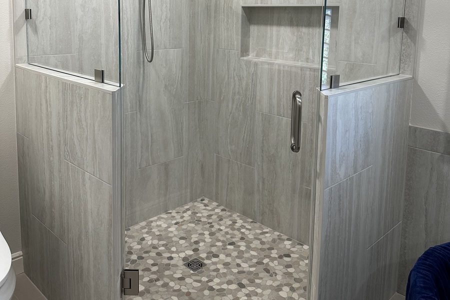 A finished neo angle shower with frameless glass, large grey porcelain wall tile, a river rock pebble floor, a recessed niche and a handheld shower on a slide bar