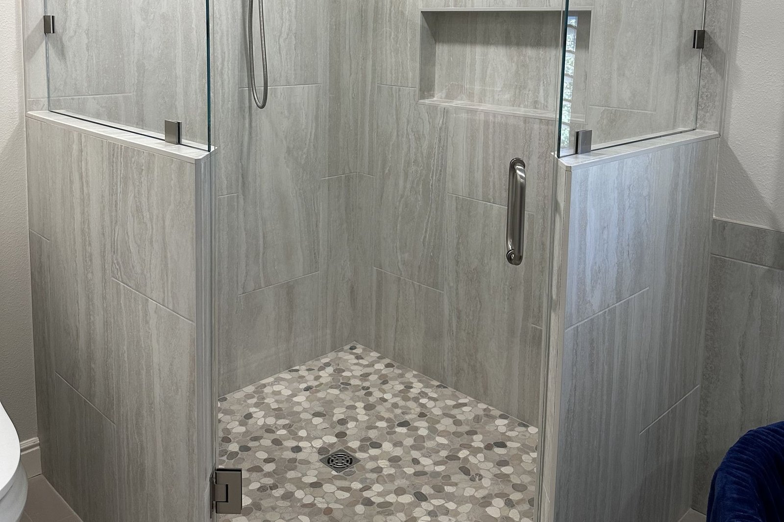 A finished neo angle shower with frameless glass, large grey porcelain wall tile, a river rock pebble floor, a recessed niche and a handheld shower on a slide bar