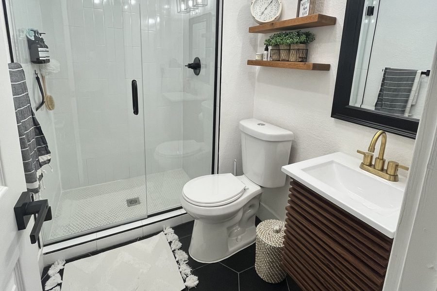 A finished small bathroom with vertically stacked white tile in the shower behind a glass panel, black hexagon floor tile, a fluted wood vanity with a brass faucet and a black framed mirror