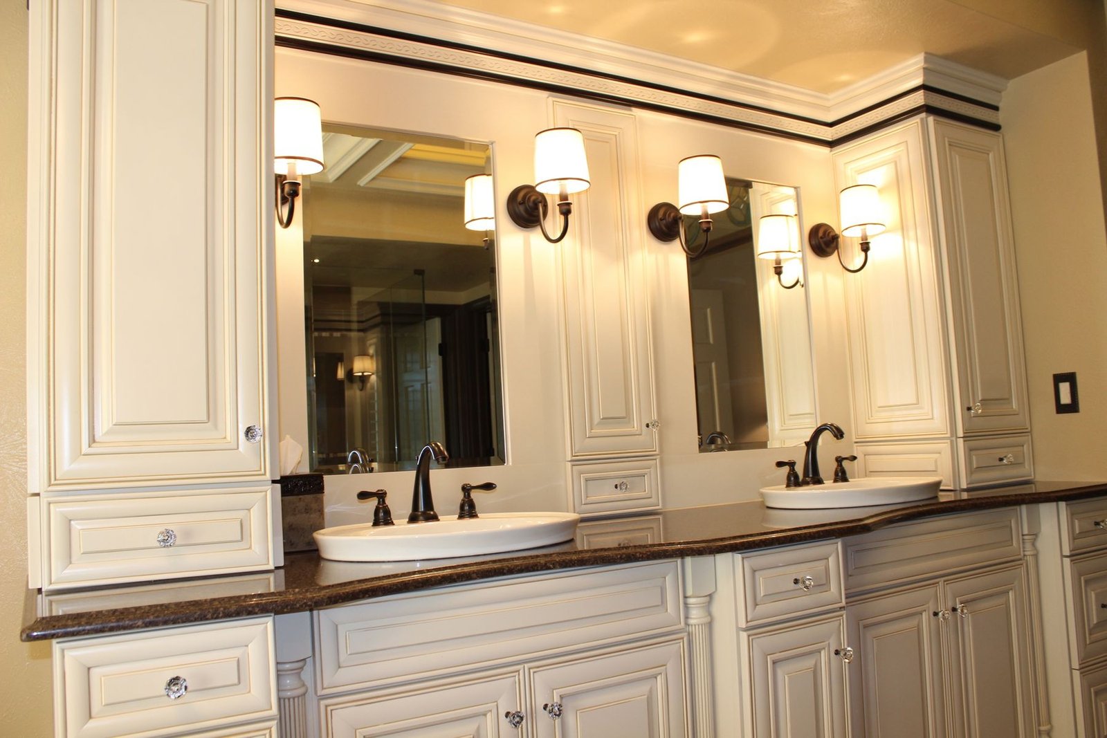 A finished bathroom with antique white glazed cabinetry, fluted pilaster details, a dark granite counter, two sinks, oil rubbed bronze faucets and sconces mounted directly on a full width mirror