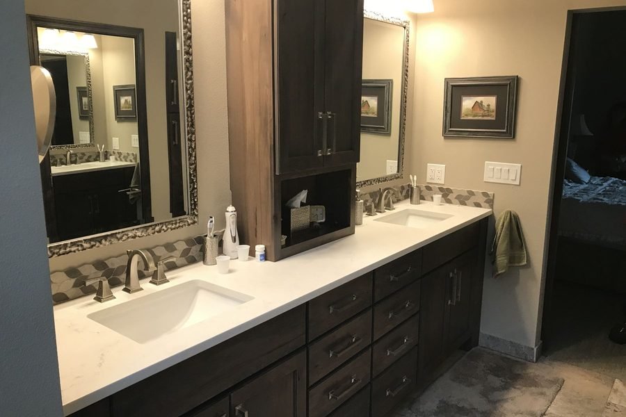 A finished bathroom vanity in dark stained cabinets with a white counter, two undermount sinks, a tall storage tower between them, framed mirrors and a mosaic tile band along the wall