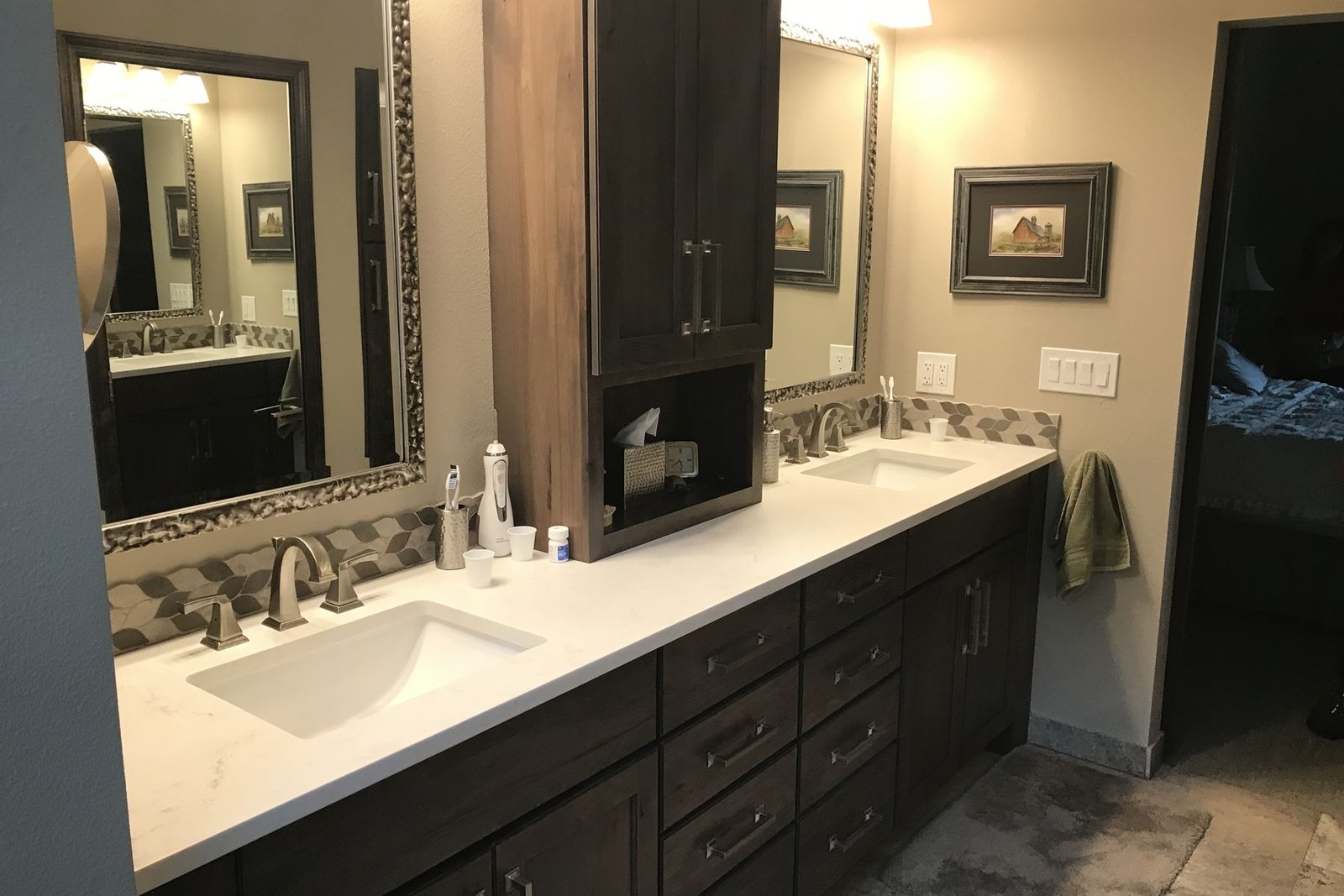 A finished bathroom vanity in dark stained cabinets with a white counter, two undermount sinks, a tall storage tower between them, framed mirrors and a mosaic tile band along the wall