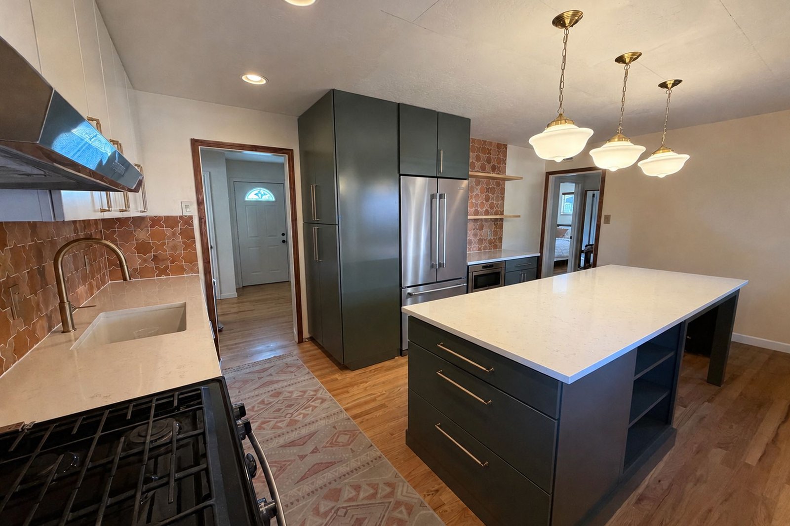 The finished kitchen seen from the range end: a white quartz island under three brass schoolhouse pendants, a sink with a brass faucet below a terracotta tile backsplash, and deep green cabinetry running back to a full-height pantry with a stainless refrigerator