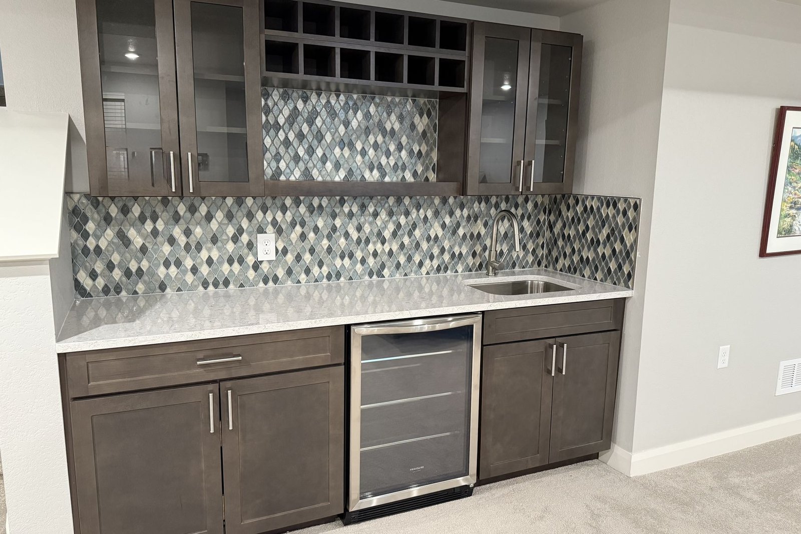 A finished wet bar with grey brown stained cabinets, glass fronted upper doors, a central wine cubby rack, a white counter with an undermount sink, an undercounter beverage refrigerator and a blue and grey harlequin mosaic backsplash
