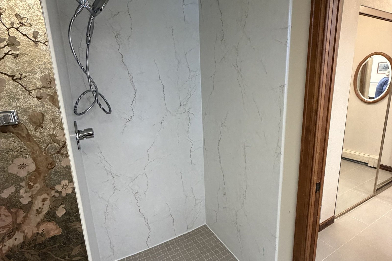 A finished shower lined in white marble-look laminate panels with no grout lines anywhere on the walls, with a chrome handheld shower head and a small-tile shower pan, opening onto a hallway with pale tile flooring