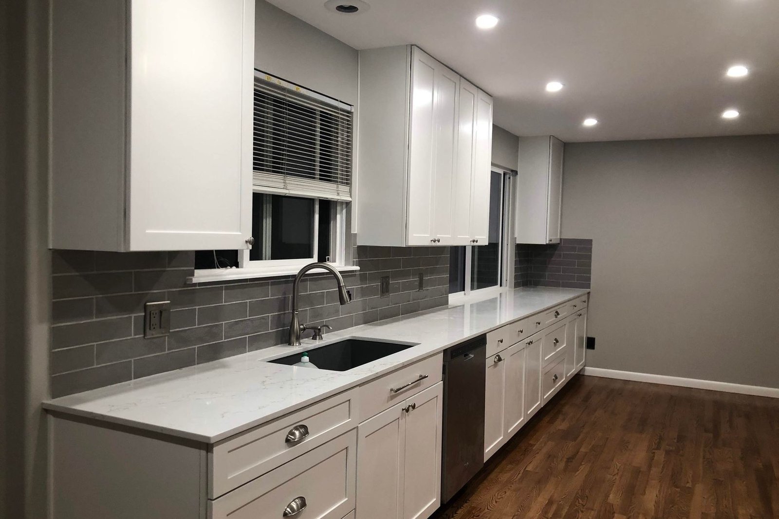 A finished galley kitchen with white shaker cabinets along one wall, quartz counters, a grey subway tile backsplash, a black undermount sink and faucet, and a dark wood floor