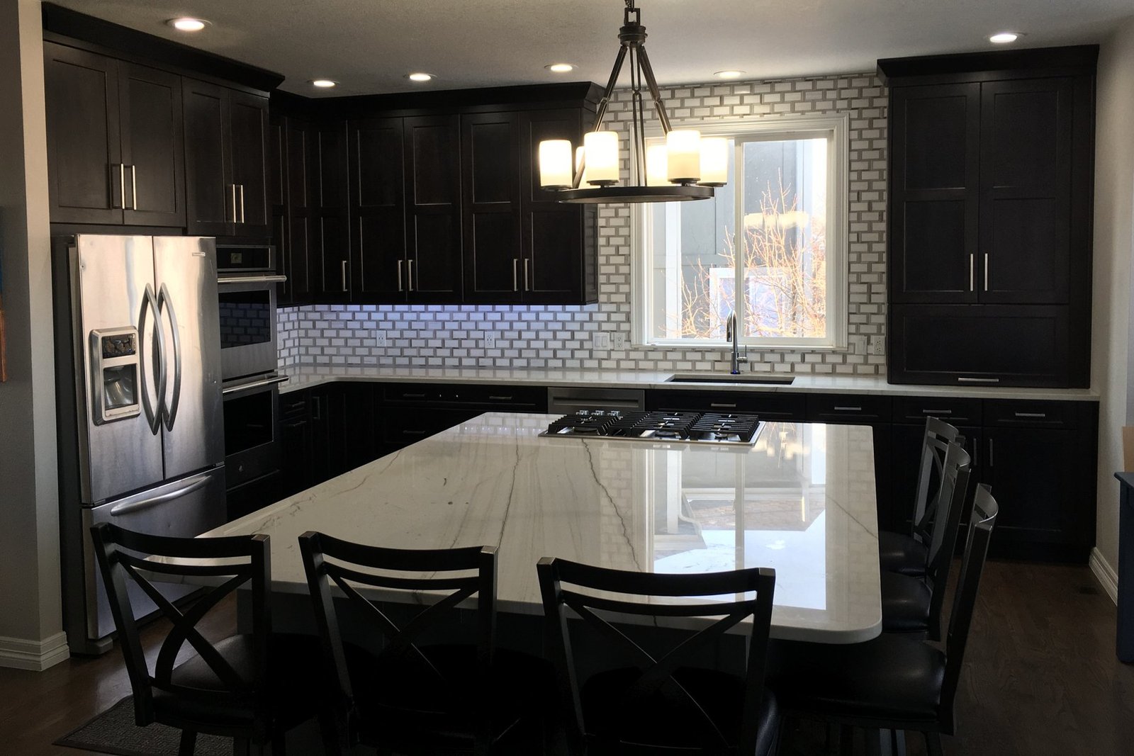 A finished kitchen with espresso shaker cabinets running to the ceiling, a long white stone island with a gas cooktop and seating, a brick-set tile backsplash, stainless wall ovens and a chandelier over the island