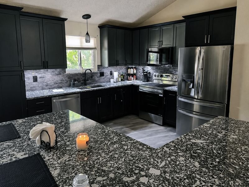 The same kitchen after, seen across the island: charcoal shaker cabinets, speckled granite counters, a stone-look tile backsplash, stainless appliances and a window over the sink under a vaulted ceiling