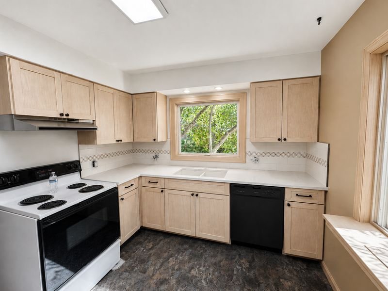 The same kitchen after: the original cabinets refaced with light full overlay doors, a pale countertop and new flooring at the same window over the sink