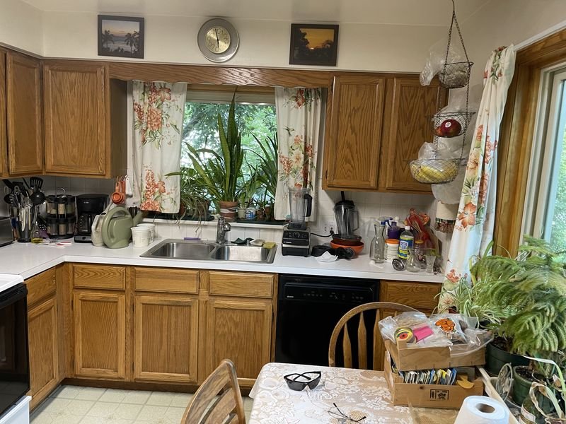 Kitchen before: the original cabinets with oak doors and laminate counters, with floral curtains at the window over the sink