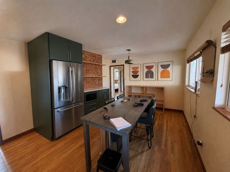 The same wall after: a full-height deep green pantry with a built-in stainless refrigerator, a counter with a microwave below it, and open wood shelves on terracotta tile