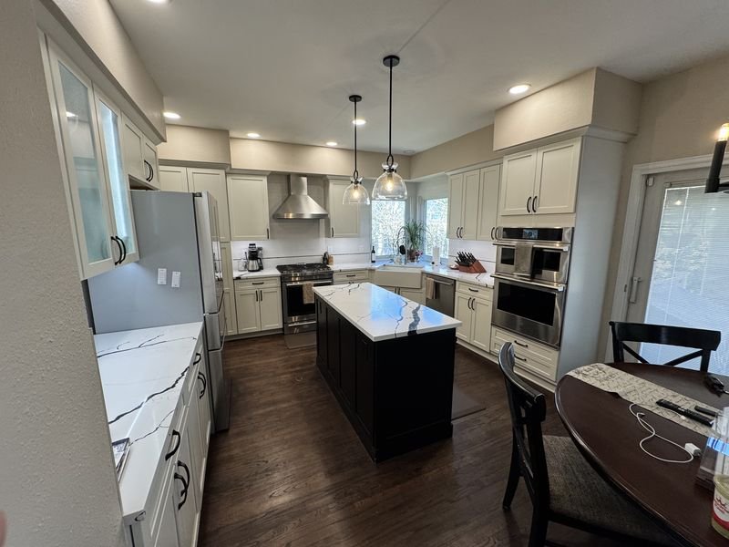 The same kitchen after: light perimeter cabinets, a dark island with pendant lights and a stainless chimney hood