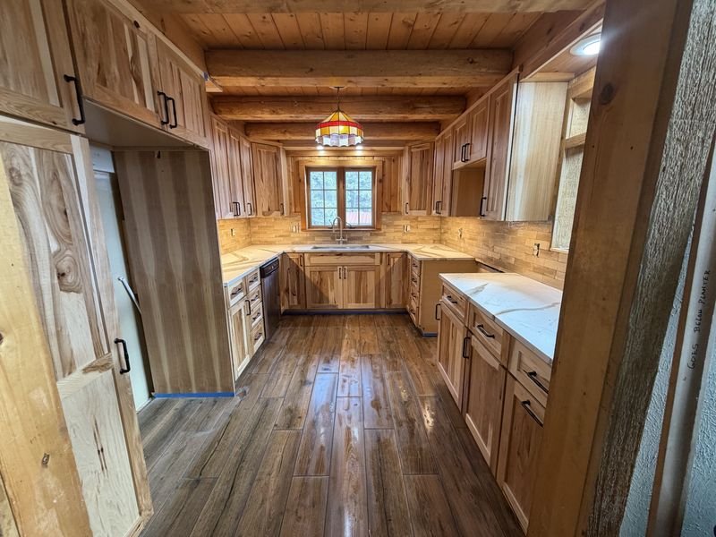 The same kitchen after: hickory shaker cabinets with black pulls, white veined counters, a brick-style tile backsplash and a wood-look plank floor under the same log beams and pendant