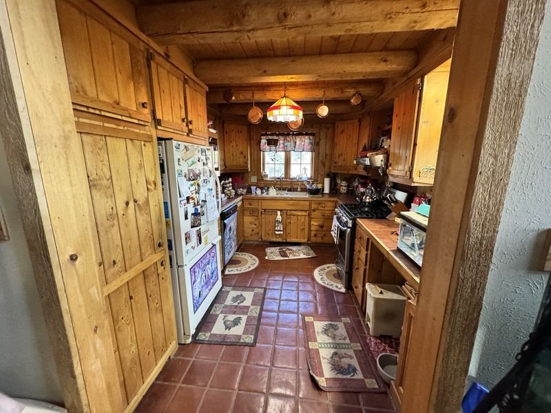 Log-home kitchen before: knotty pine plank cabinets, a white refrigerator, tiled counters and a red square-tile floor under log beams and a stained-glass pendant
