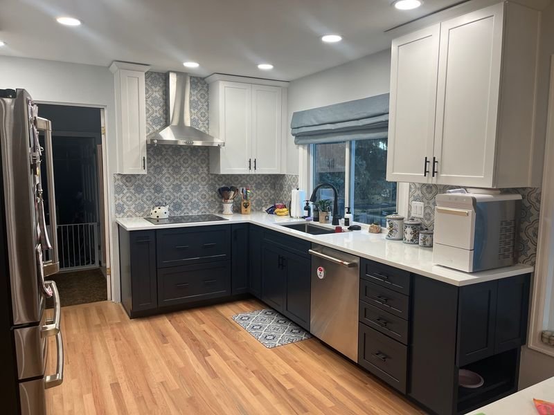 The same window wall after: white upper cabinets over navy bases, patterned tile backsplash and a stainless chimney hood