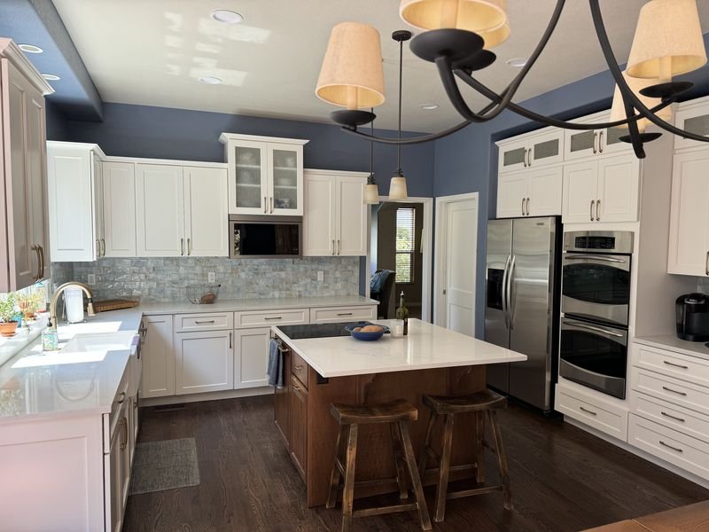 The same kitchen after: white shaker cabinets, white quartz counters, a light stone backsplash and blue walls, with the wood island kept and re-topped in white
