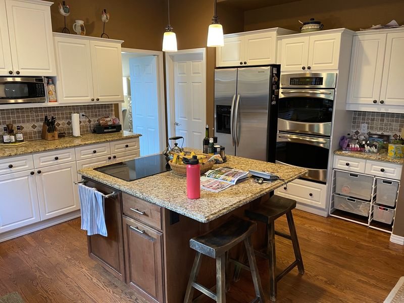Kitchen before: white raised-panel cabinets with speckled granite counters, a tan tile backsplash and brown walls, and a dark wood island under two pendant lights