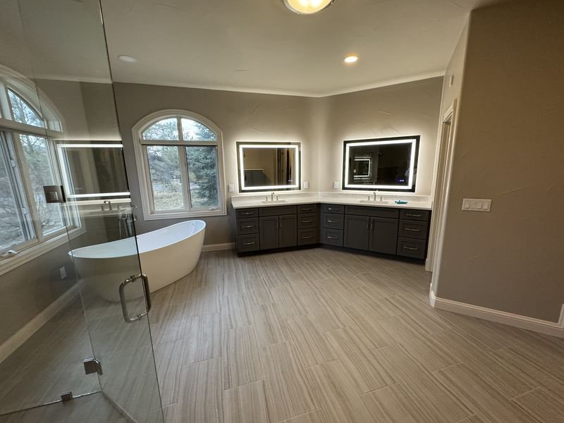 The same primary bathroom after: a freestanding tub beneath arched windows, dark vanities with lit mirrors and plank tile flooring