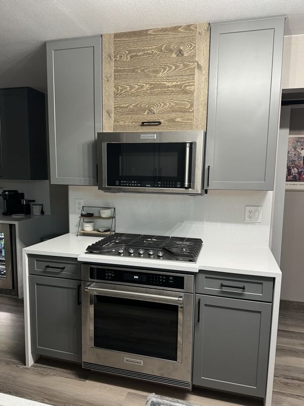 The same range wall after: gray shaker cabinets, a wood-faced panel above a stainless microwave, a gas cooktop in a white quartz counter with a stainless wall oven below