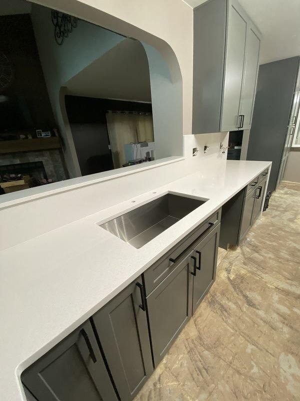 The same sink run after: gray shaker cabinets and a single deep undermount sink in a white quartz counter that continues to the pass-through ledge