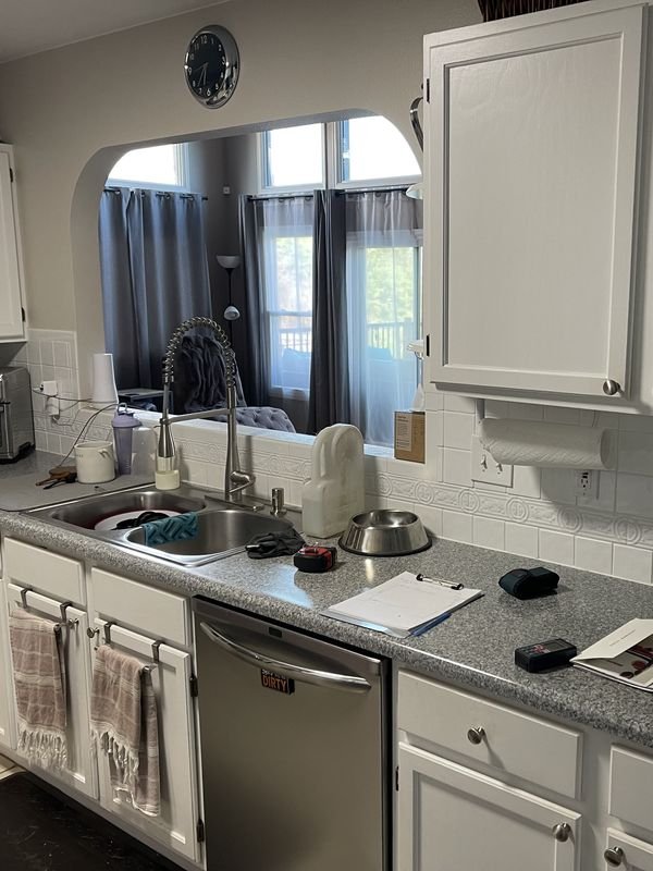 Kitchen sink run before: white cabinets, a drop-in stainless double sink in a laminate counter and an arched pass-through to the living room