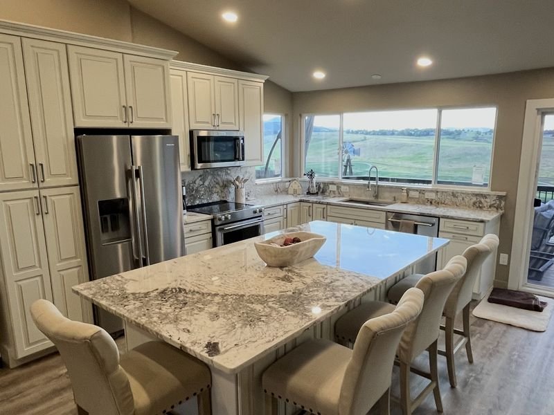 The same kitchen after: cream raised-panel cabinetry, a granite island with seating for five, and windows over the sink looking out to open country