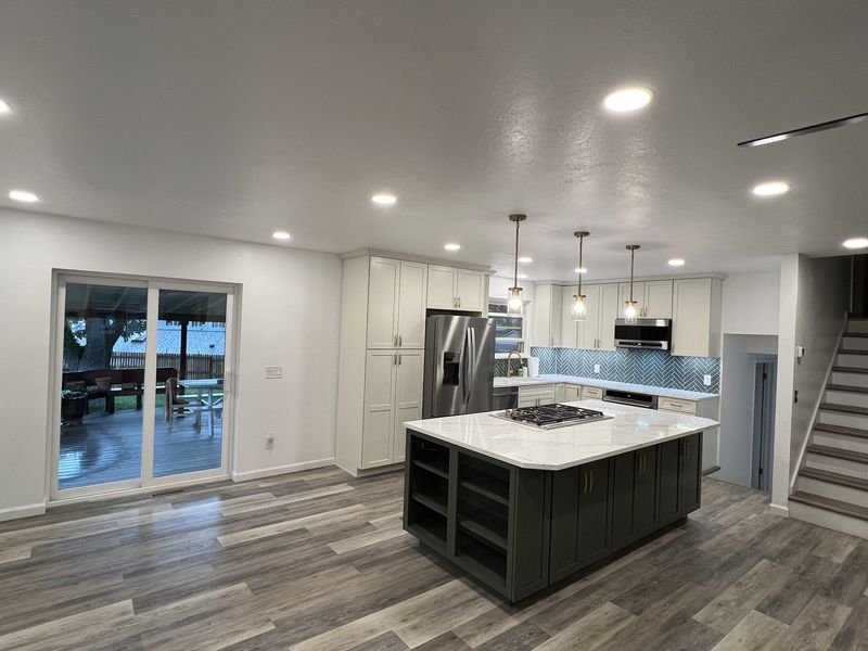 The same space with the walls out: white perimeter cabinets, a deep green island with open shelving and a gas cooktop, a herringbone tile backsplash, and a patio slider where the dining room window had been
