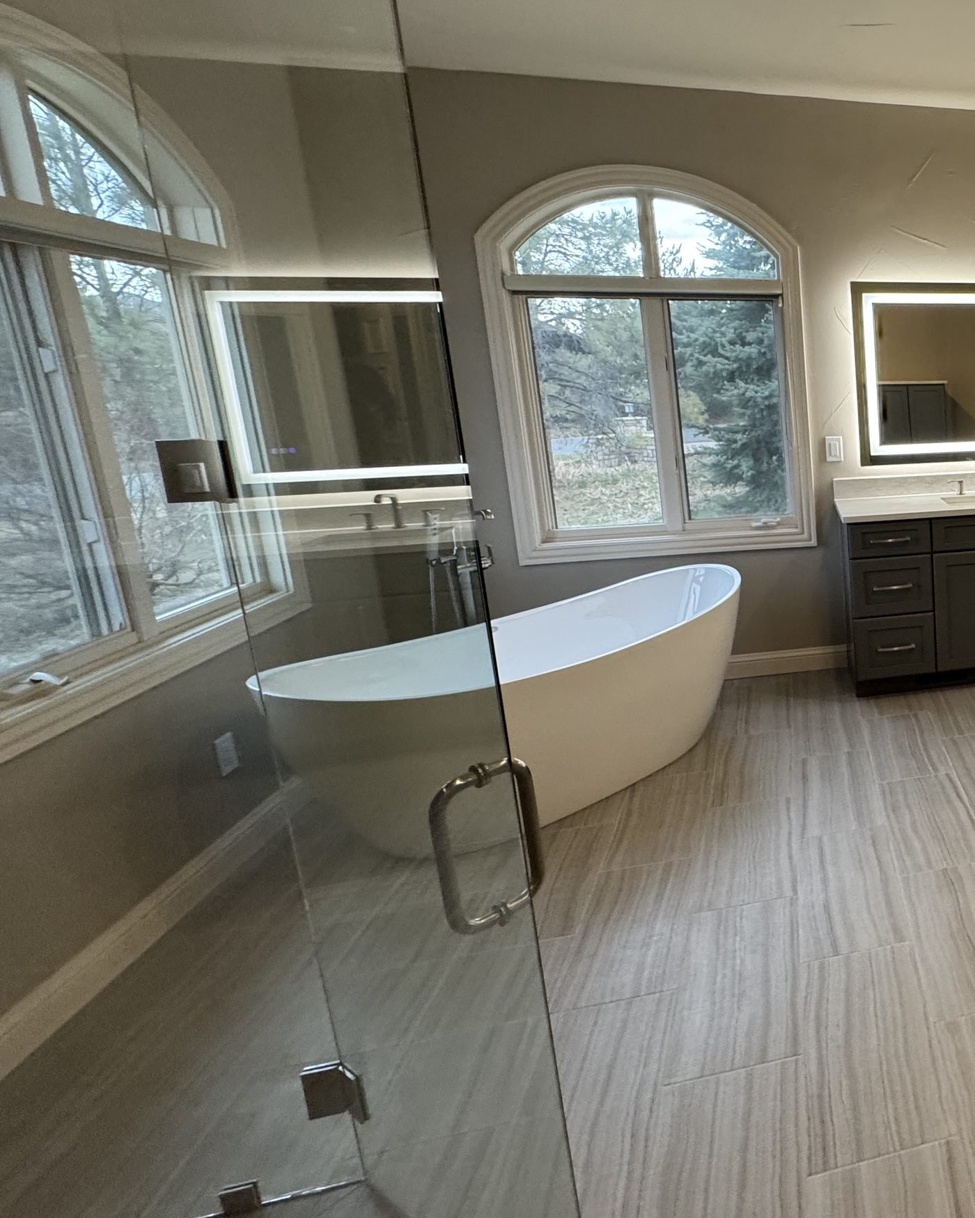A white freestanding soaking tub beneath an arched window in a primary bathroom, beside a glass shower panel, on plank tile flooring