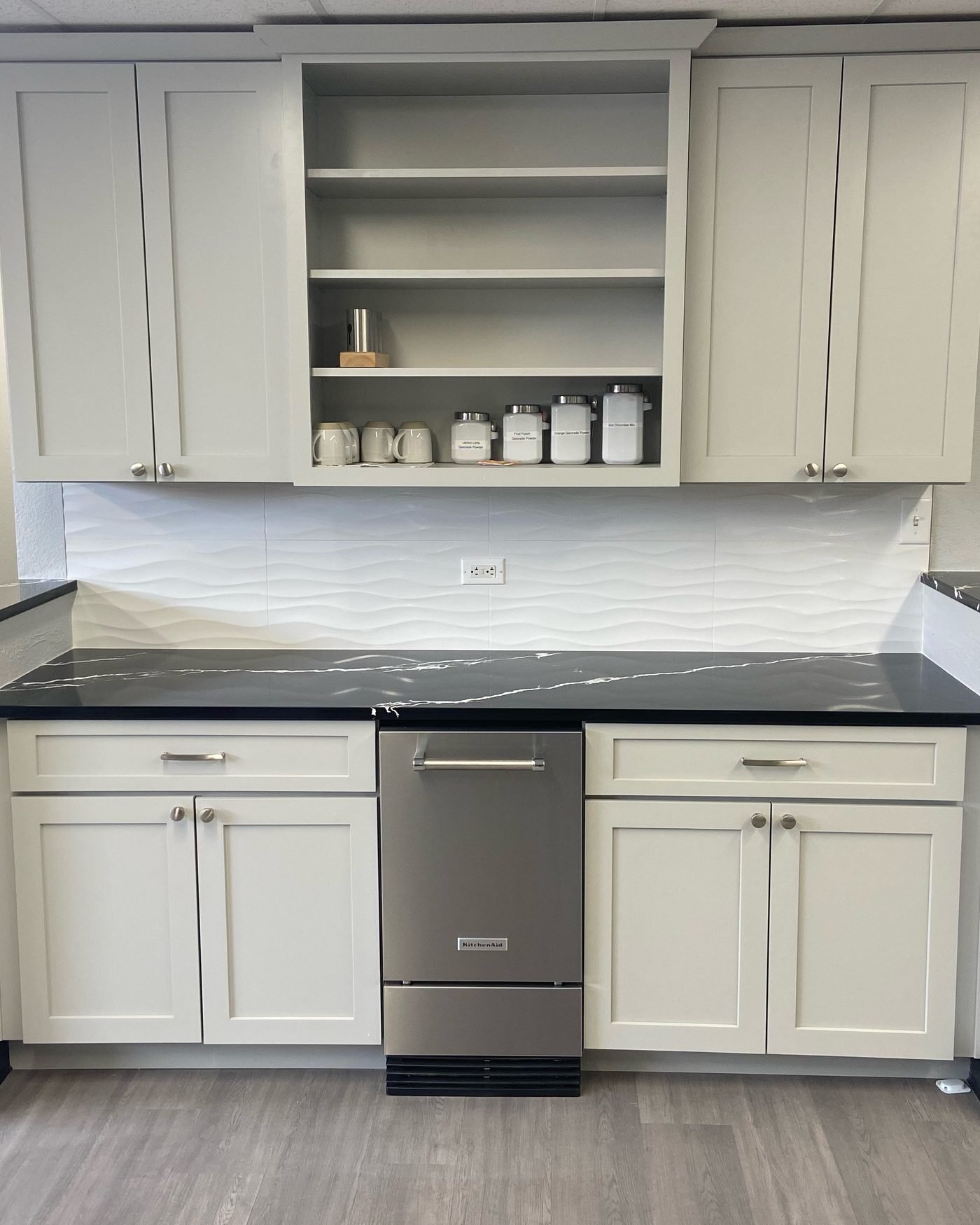 A run of pale grey shaker cabinets under an open shelf of canisters and mugs, with a black stone counter and a white wave-textured tile backsplash
