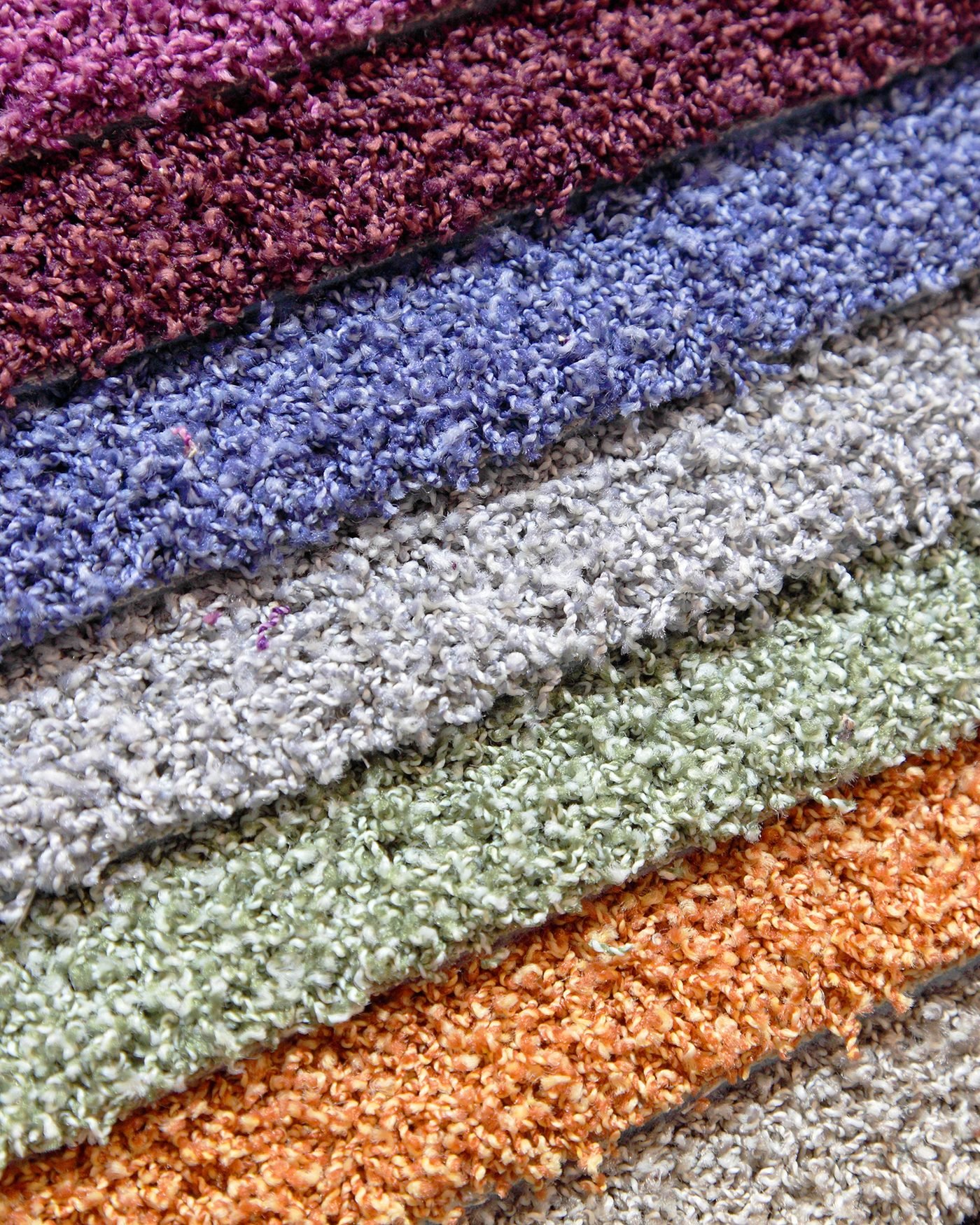 Six carpet samples stacked in a fan, in plum, periwinkle, grey, sage, orange and taupe, each showing its own cut pile texture along the exposed edge