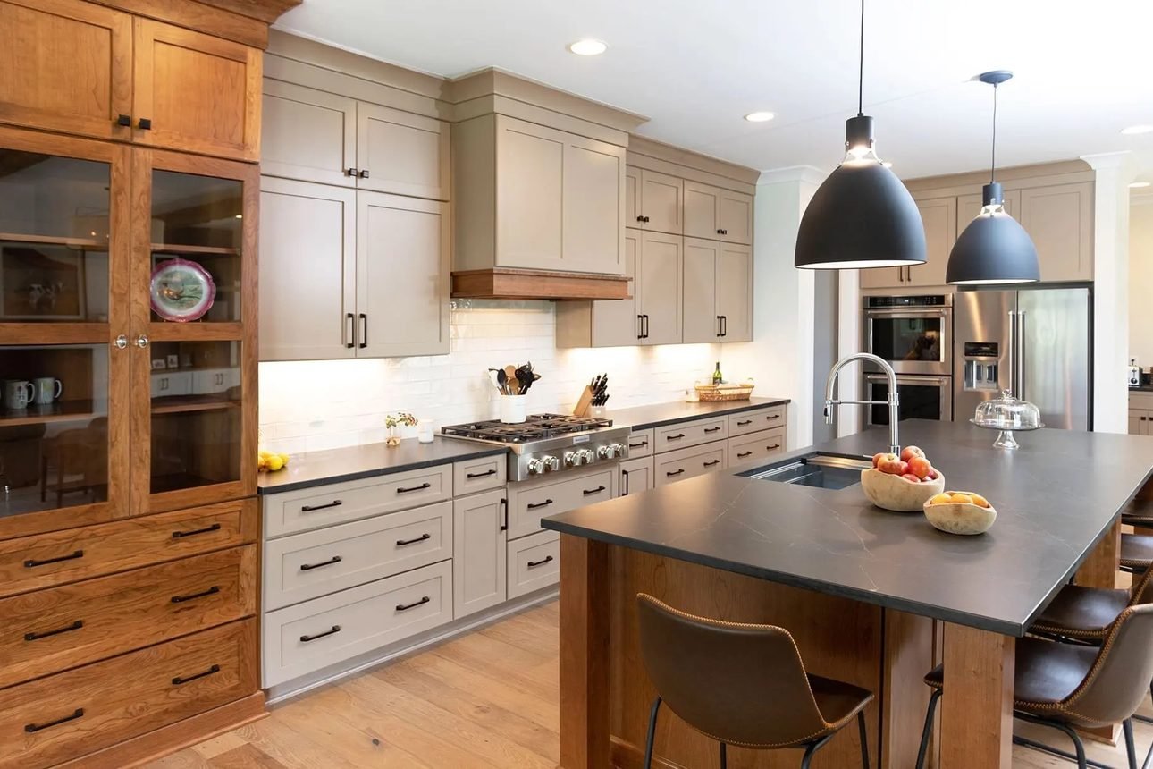 A kitchen in taupe painted cabinetry with a cherry glass-front hutch, dark stone counters, a wood chimney hood and two black dome pendants over a wood island