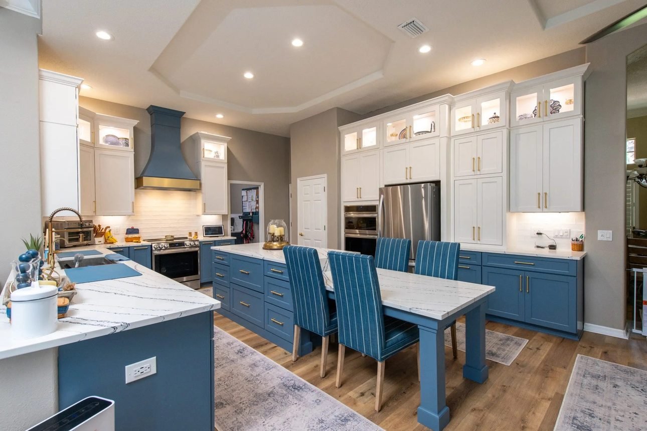 A kitchen in white upper cabinets and blue lowers with brass hardware, a blue chimney hood, lit glass display cabinets and a matching blue table with striped chairs