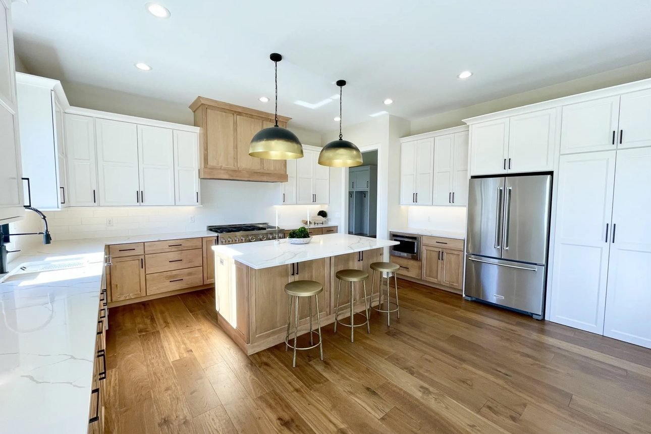 A kitchen in white uppers with natural oak lower cabinets and island, a wood chimney hood, brass dome pendants and wide plank oak flooring