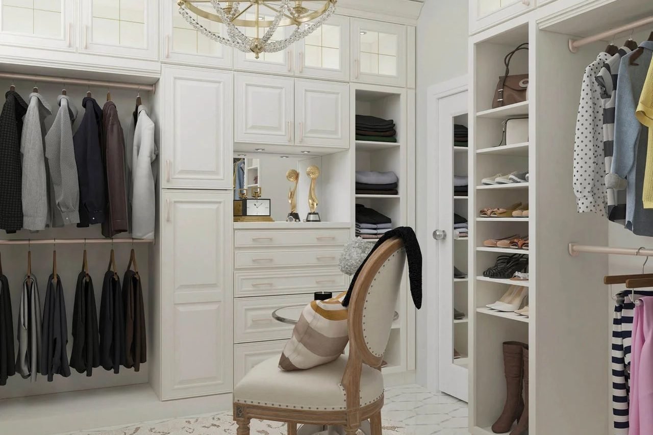 A white fitted walk in closet with a brass chandelier, hanging rails, glass front drawers and open shoe shelving, around an upholstered chair