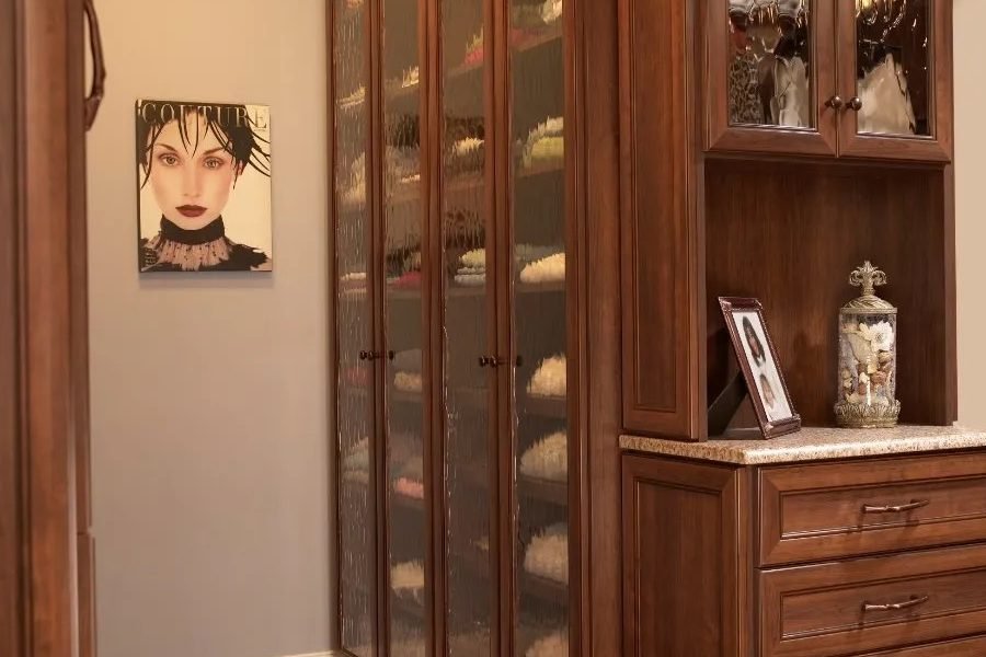 A dressing room in dark stained cabinetry with glass fronted display cabinets, drawer banks and a framed portrait on the end wall