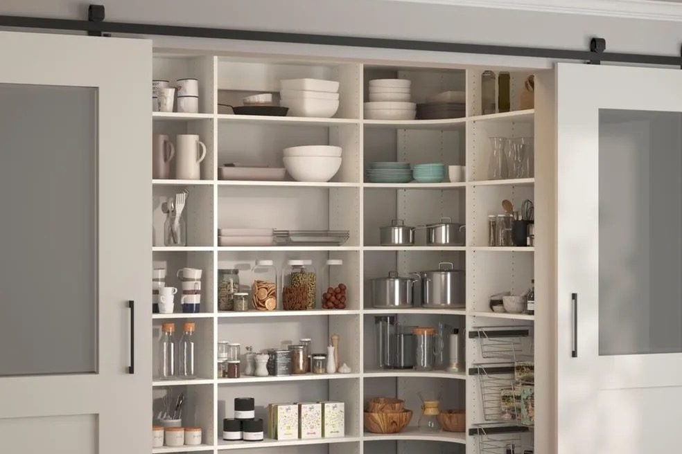 A corner pantry in white shelving stocked with dishes, jars and canisters, behind a pair of sliding barn doors on a black rail