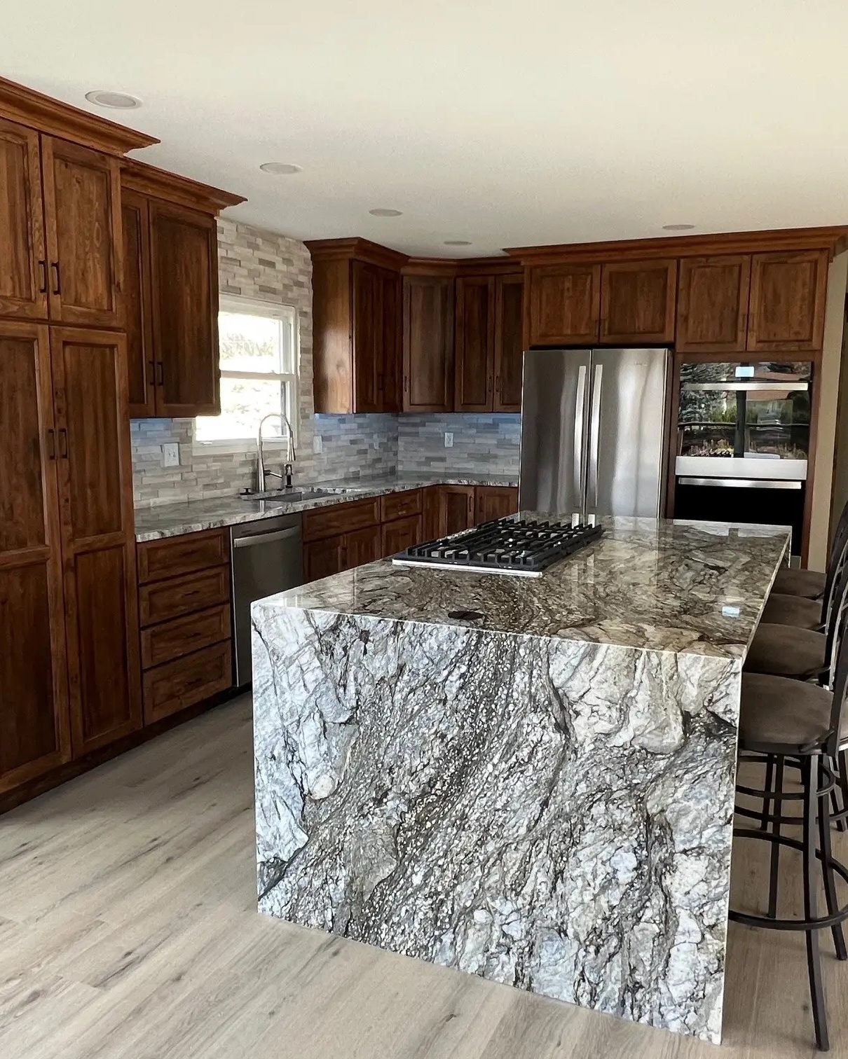 A kitchen island in heavily veined grey and brown granite with a waterfall edge, in front of knotty alder cabinetry, a stacked stone backsplash and a stainless refrigerator