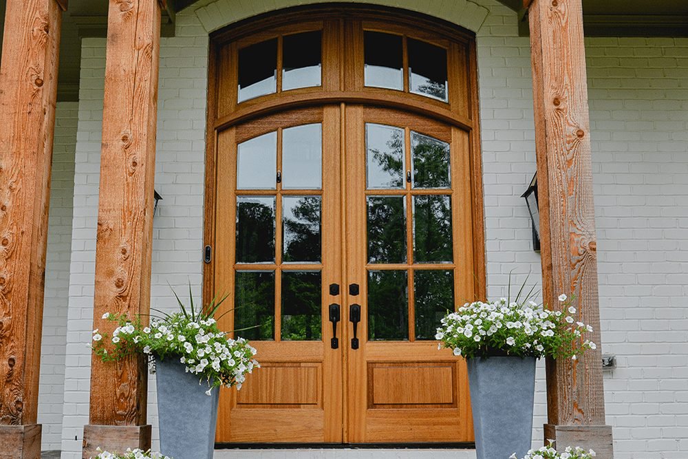 A pair of arched wood entry doors with divided glass under a covered porch, flanked by timber posts and tall planters of white flowers